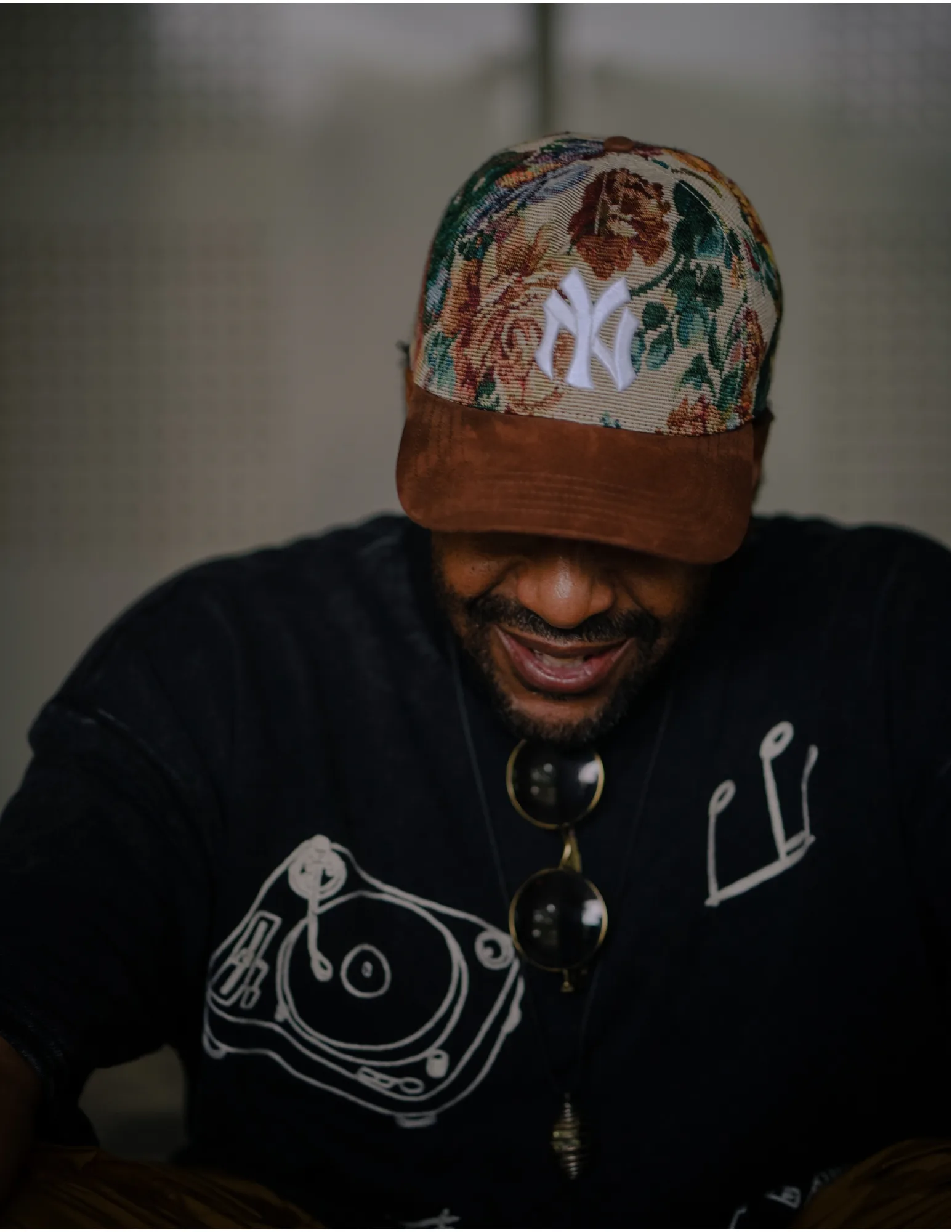 DJ P.U.D.G.E. wearing a black jacket and baseball hat in front of a grey wall. Face is obscured.