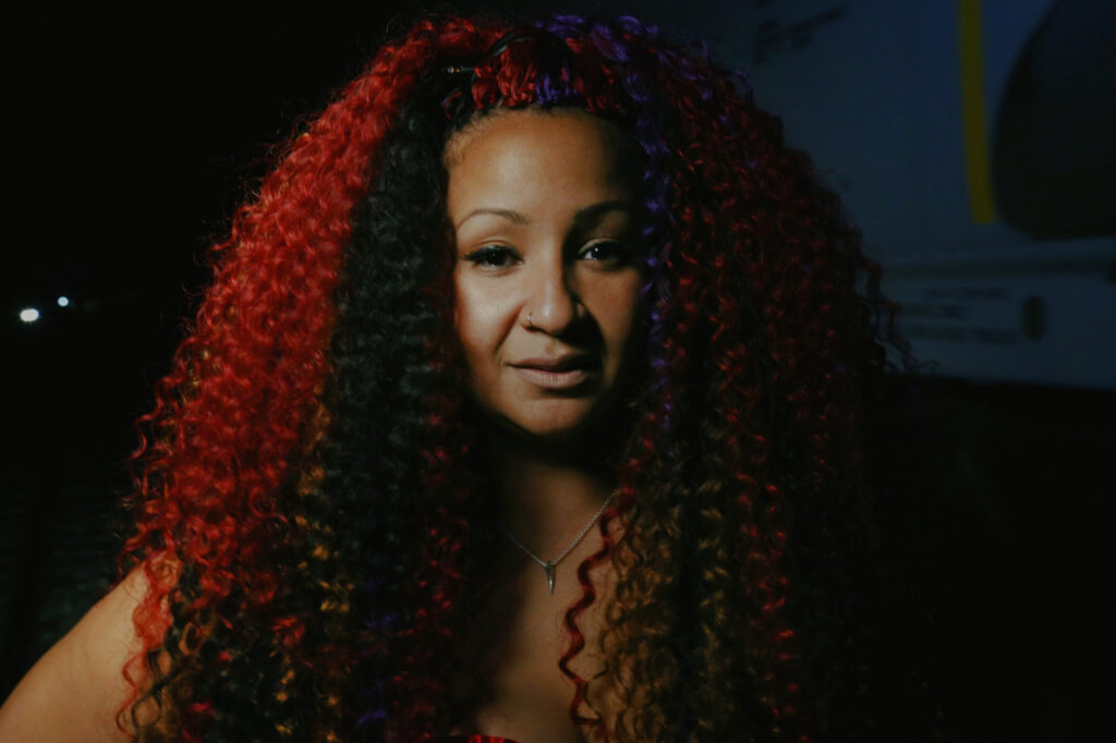 Woman with long, curly hair and a necklace, possibly in a dimly lit room.