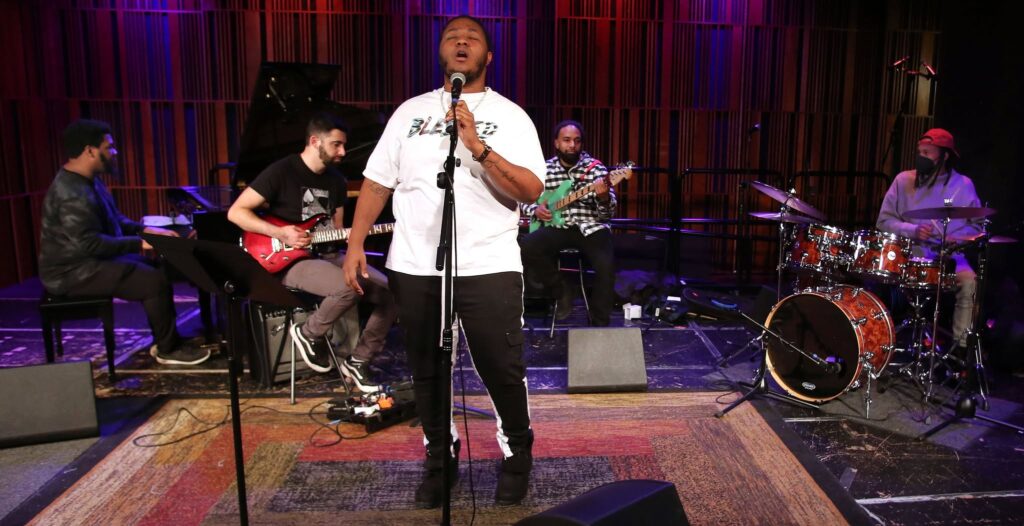 A man singing into a microphone on a stage, with a guitarist and drummer in the background. The man is wearing a white t-shirt and black pants, and has short hair. He is holding a microphone stand and appears to be singing into it. Behind him, to the left, is a man playing an electric guitar, and to the right is a man playing drums. The background is a stage with a red curtain and a drum set.