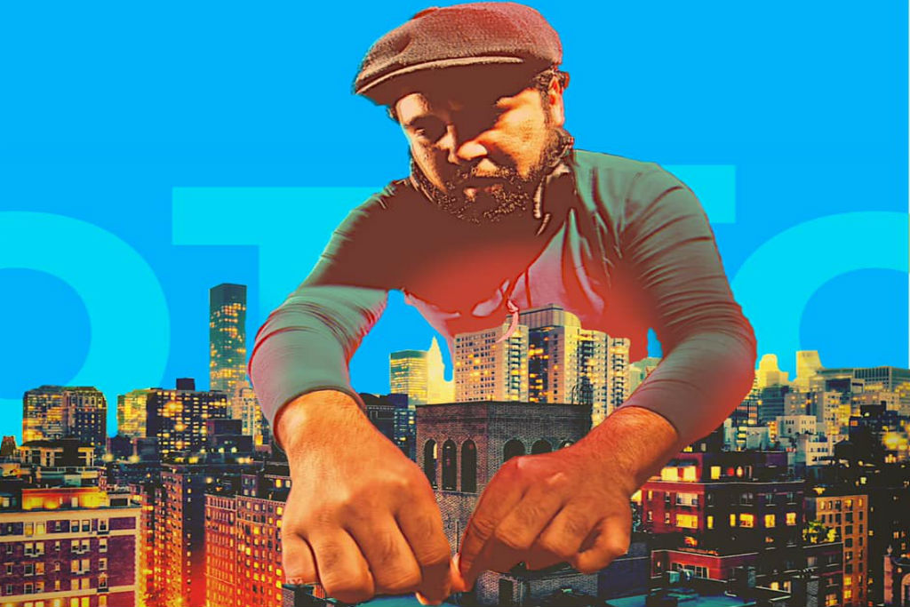 A man in a hat and long-sleeved shirt, reaching out over a cityscape at night.