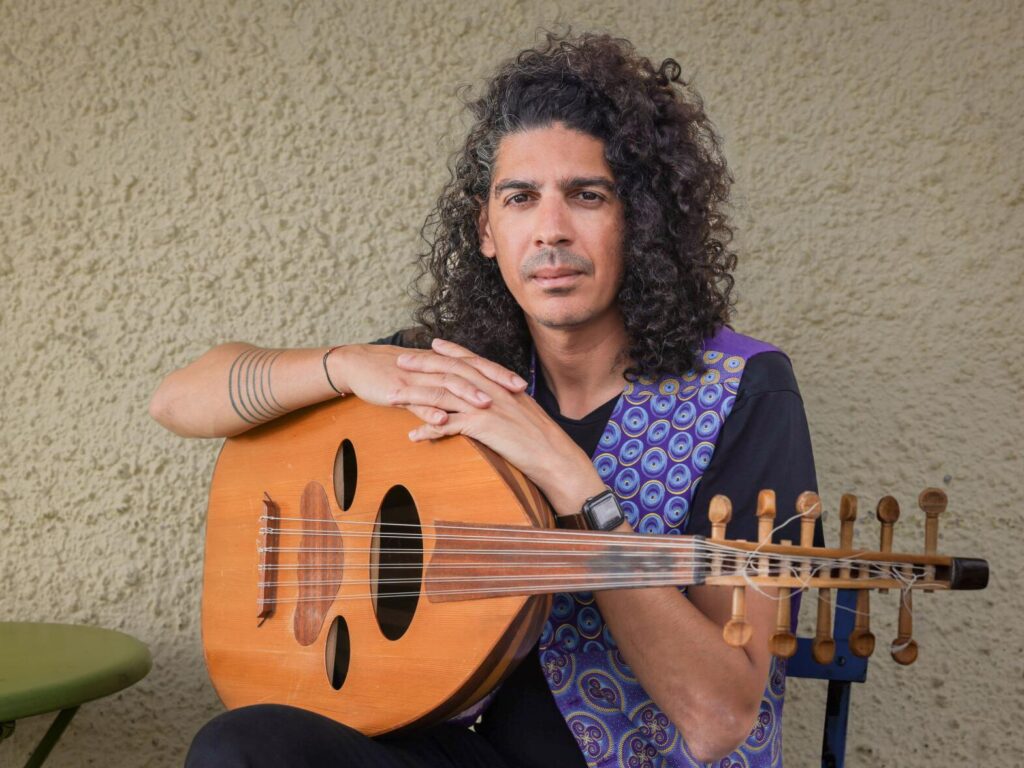 A man with curly hair sits in a chair, holding a stringed instrument. The instrument has a round body and a long neck with tuning pegs. The man wears a blue patterned vest over a black shirt and black pants. He has a watch on his left wrist and a bracelet on his right wrist. The background is a beige wall.