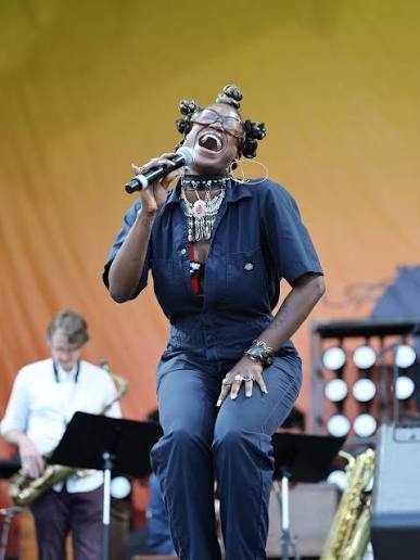 Black woman in blue overalls singing into microphone on stage, with saxophonist behind her.