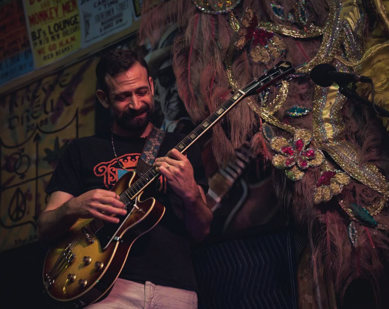 A man with dark hair and a beard, wearing a black shirt, plays a brown electric guitar in front of a wall with graffiti and a colorful mural.