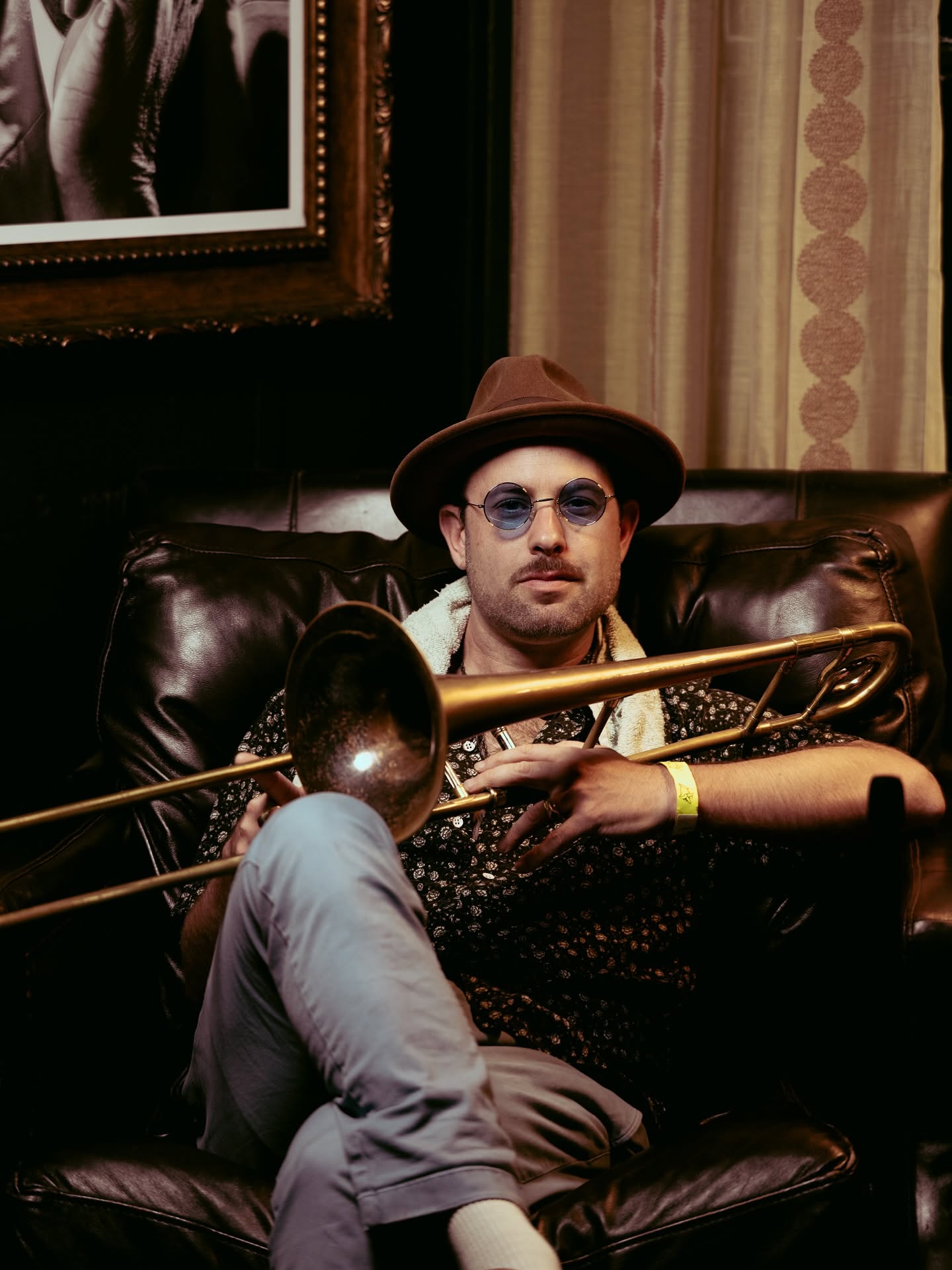 A man in a hat sits on a leather couch, holding a trumpet.