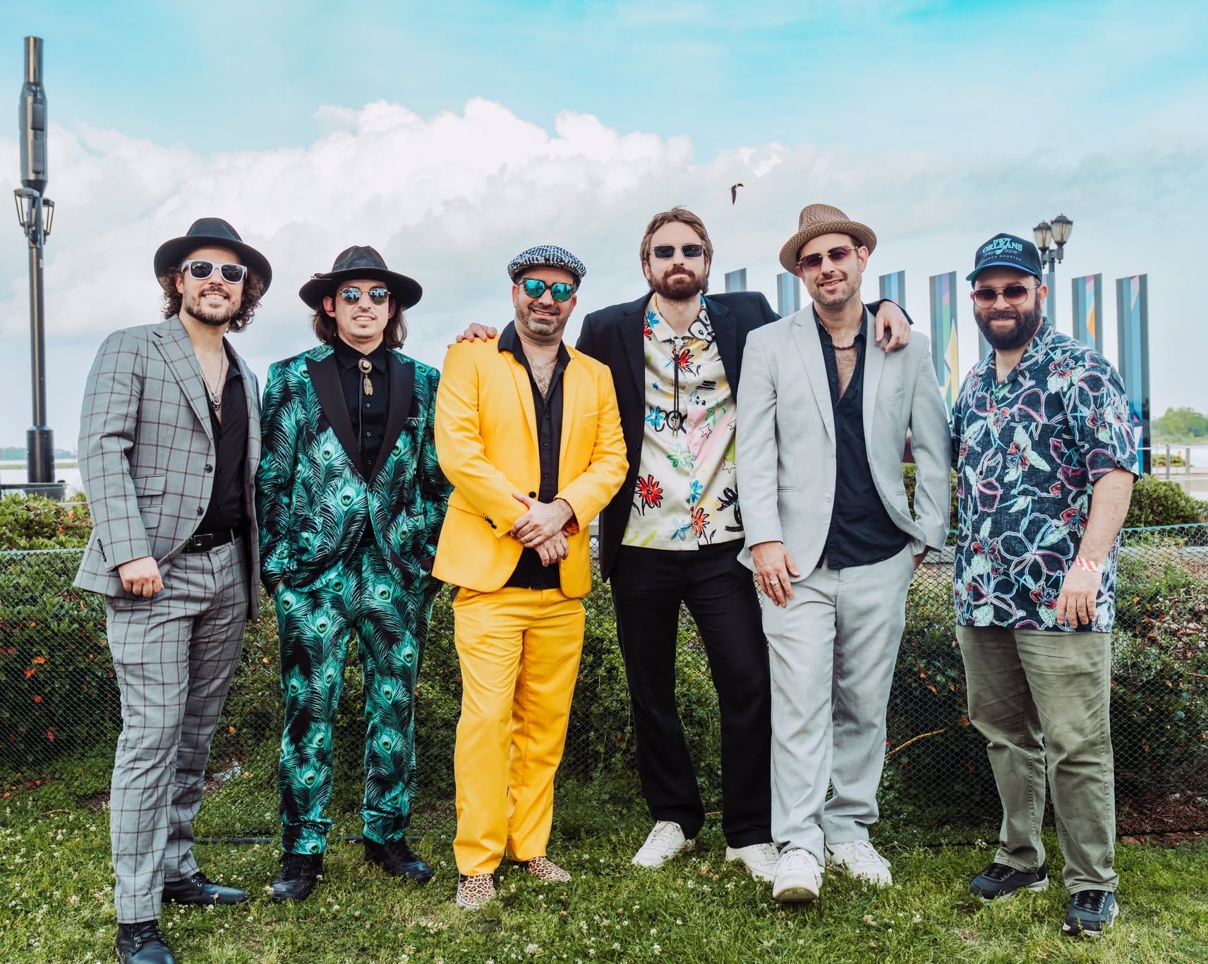 Five men standing in a line, posing for a photo. They are dressed in various outfits, including suits, hats, and sunglasses. The background is a blue sky with white clouds.