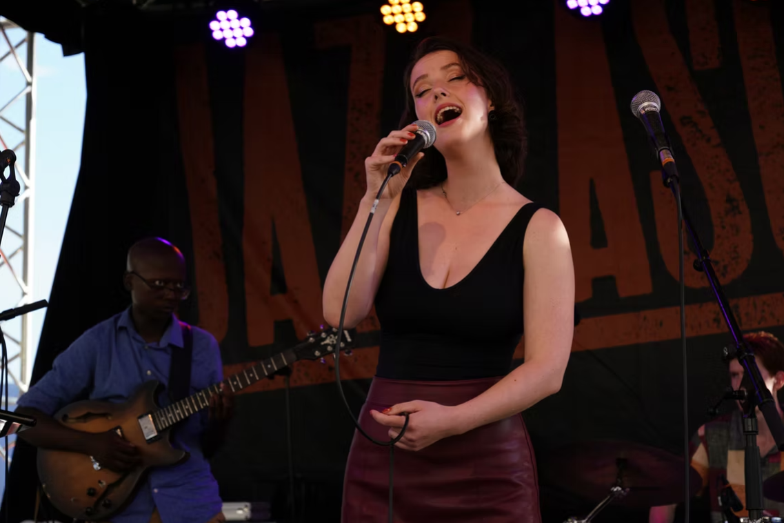 A woman singing into a microphone on stage, with a man playing guitar in the background, possibly at a music venue or event.