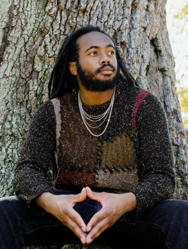 Black man with long hair and beard, wearing a sweater and silver chain, sitting in front of a tree trunk.