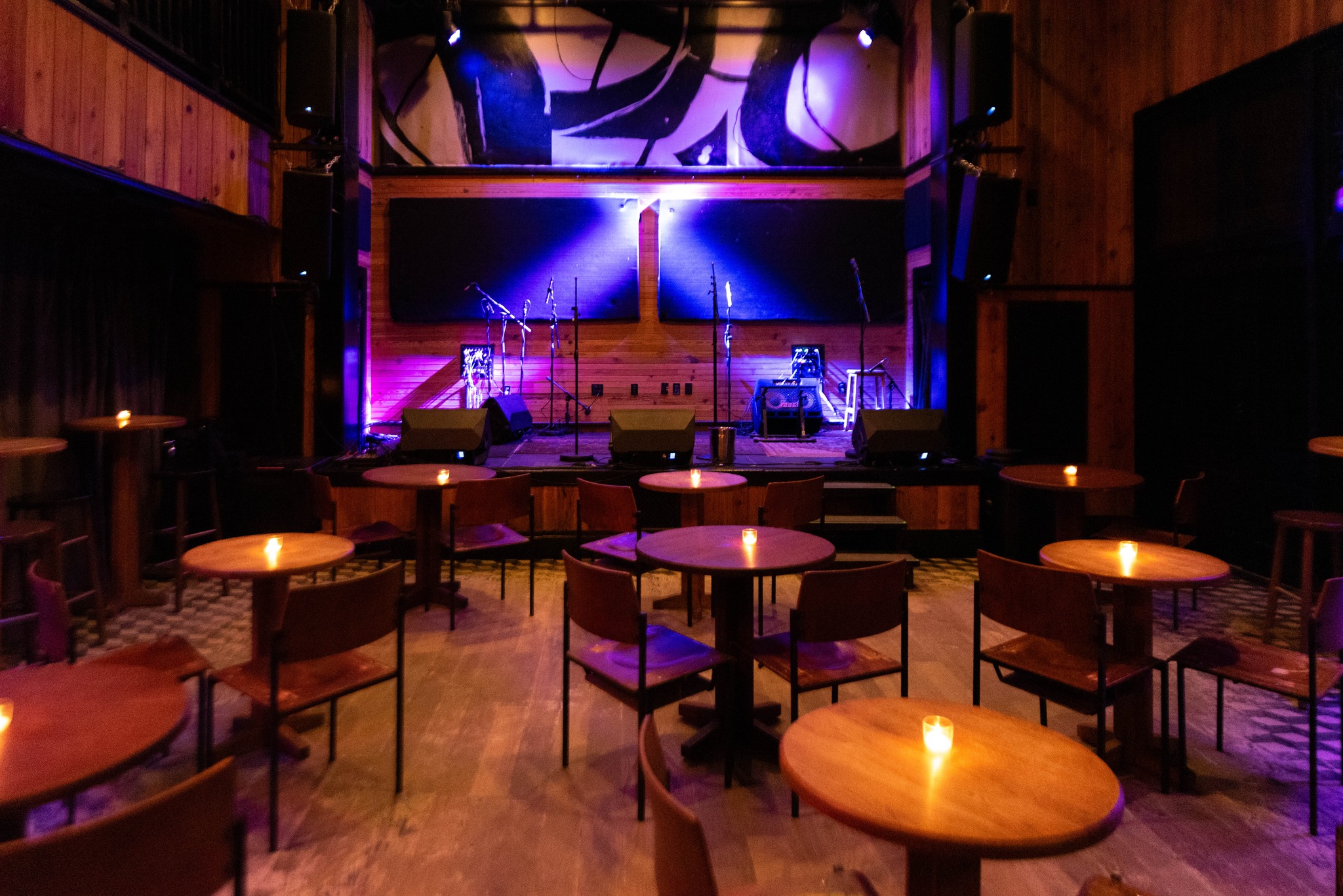 A dimly lit room with tables and chairs, featuring a stage with a microphone and a large screen or projection behind it.