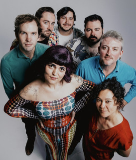 Five people posing for a photo, likely a band or group of musicians. The group consists of three men and two women, all dressed in casual attire.